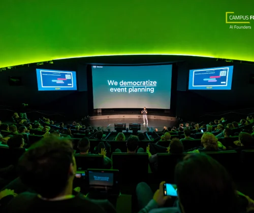 Ein Redner steht auf der Bühne in einem dunklen Hörsaal mit einem großen Bildschirm, auf dem "We democratize event planning" (Wir demokratisieren die Veranstaltungsplanung) angezeigt wird, während das Publikum zuschaut; in der oberen rechten Ecke sind das Logo von Campus Founders und eventpage.ai, eine Gästemanagement-Plattform, zu sehen. - eventpage.ai
