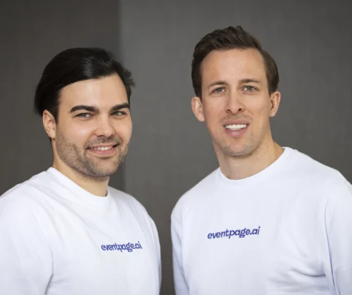 Zwei Männer in weißen Hemden mit eventpage.ai-Logo stehen nebeneinander und lächeln vor einem schlichten, dunkelgrauen Hintergrund in die Kamera, um ihre Kompetenz im Gästemanagement zu demonstrieren. - eventpage.ai
