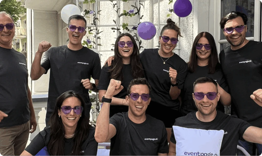 Eine Gruppe von acht Personen mit schwarzen T-Shirts und lila getönten Sonnenbrillen lächelt und reckt die Fäuste zur Feier des Tages in die Höhe. - eventpage.ai