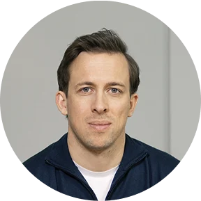 Ein Mann mit kurzen braunen Haaren und blauen Augen, der eine dunkle Jacke mit Reißverschluss über einem weißen Hemd trägt, sitzt vor einem schlichten hellgrauen Hintergrund. - eventpage.ai